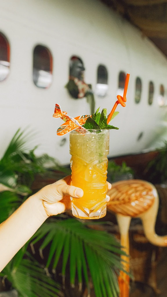 Hand holding tiki drink with paper airplane against airplane fuselage.