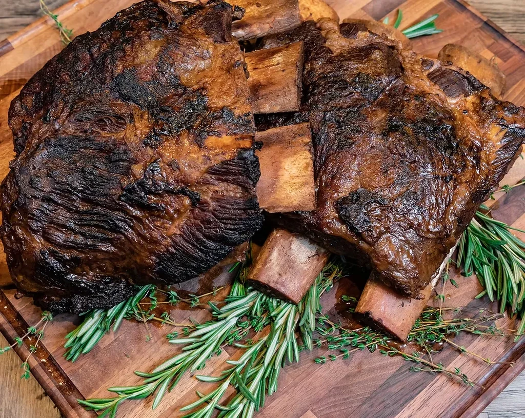 Two roasted beef short rib racks with rosemary on a wooden board.