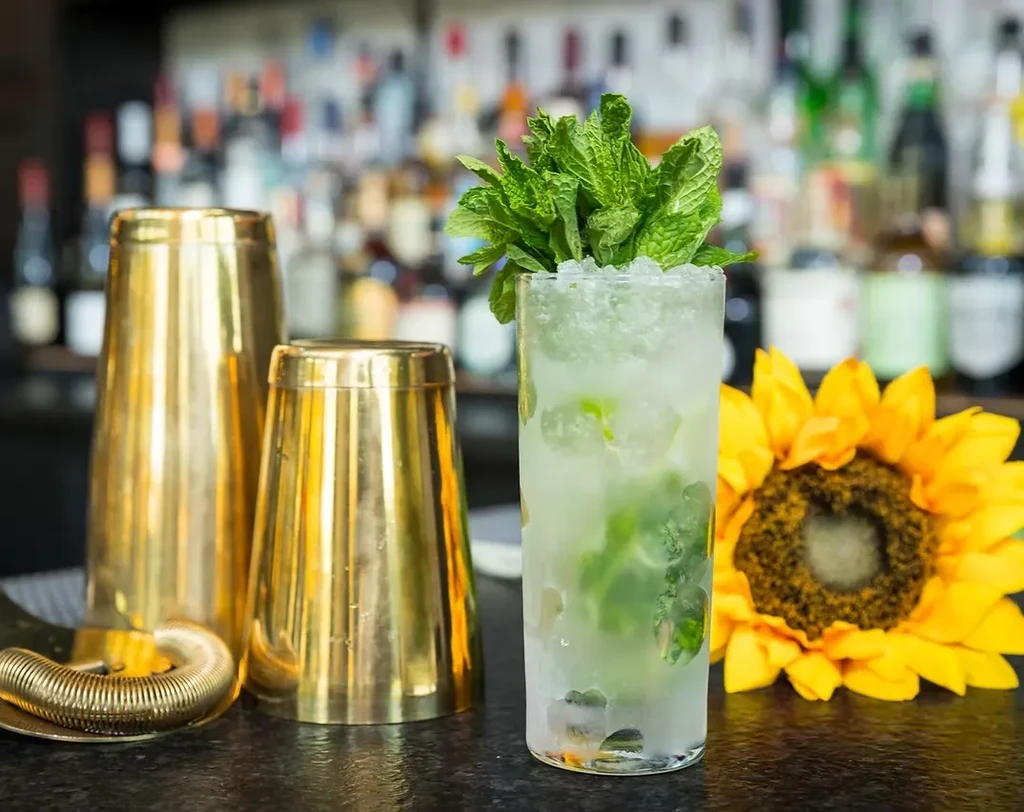 A cocktail, shakers, strainer, and sunflower on a bar top.