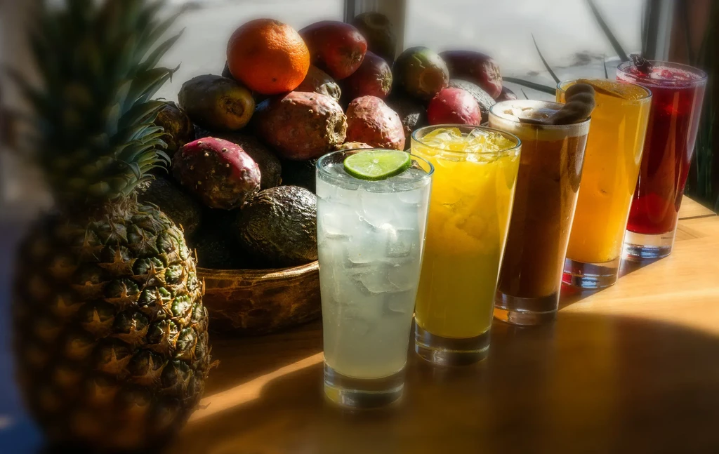 Pineapple, fruit bowl, and five colorful drinks on a sunny table.