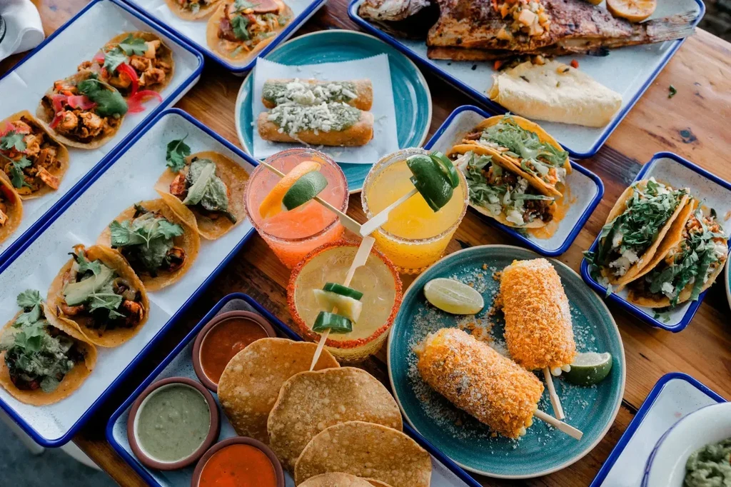 A table laden with various Mexican dishes, drinks, and garnishes.