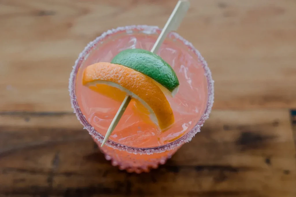 A pink cocktail with orange and lime slices on a wooden table.