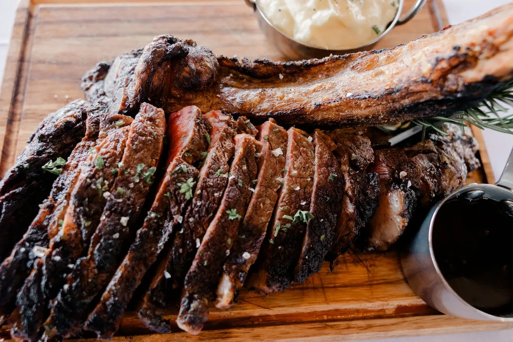 Sliced medium-rare steak with bone, sauce, and mashed potatoes on a board.