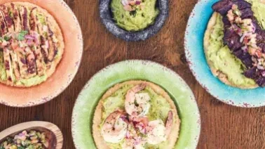 Overhead shot of five avocado tostadas with different toppings on wooden table.