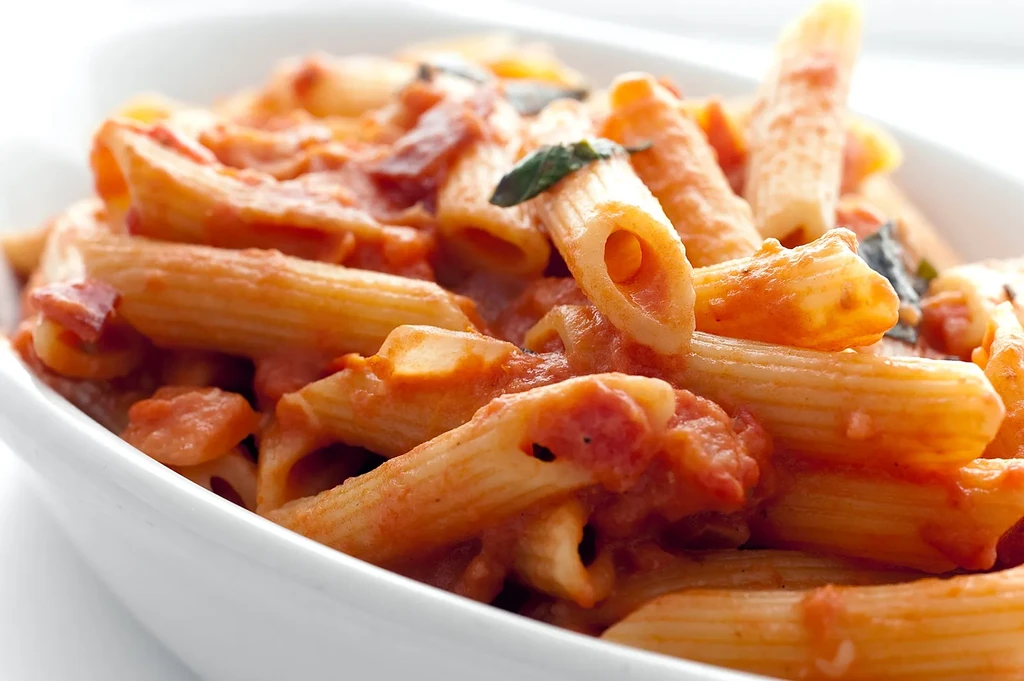 Penne pasta with tomato sauce, bacon, and basil in a white bowl.