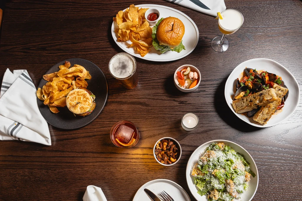 Overhead of a dining table with burgers, mussels, salads, cocktails, and beer.
