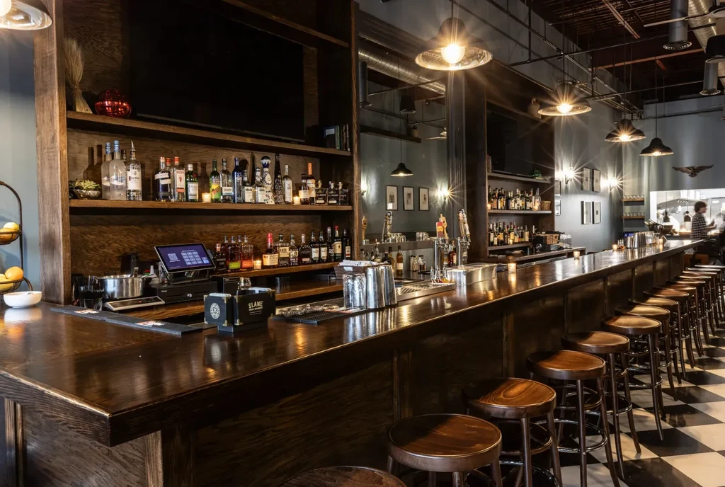 Long, dark wood bar, fully stocked with liquor, checkered floor.
