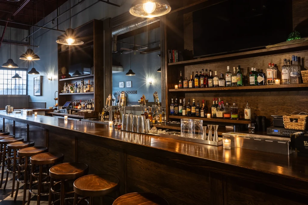 Warmly lit, industrial-chic bar with polished wood counter and liquor bottles.