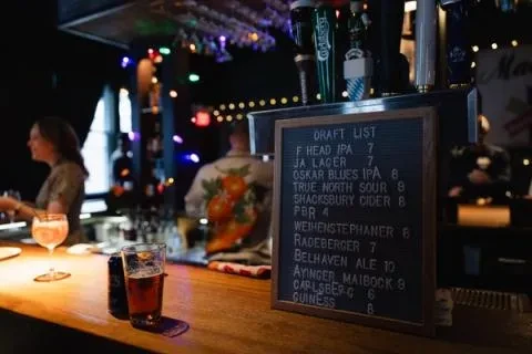 Dimly lit bar interior with drinks, 'DRAFT LIST' board, and blurred patrons.