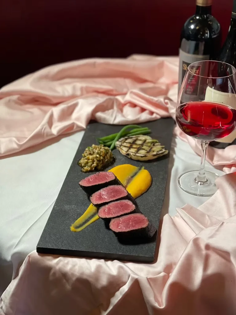 Seared beef, vegetables, and red wine on a pink tablecloth.