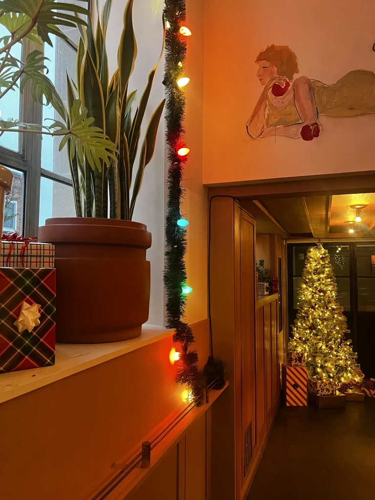 Warm interior with Christmas lights, tree, gifts, plants, and a mural.