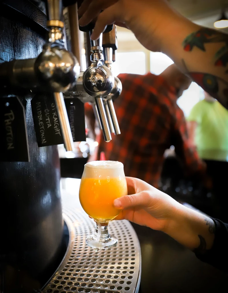 Tattooed hands pour hazy orange beer from shiny chrome taps.