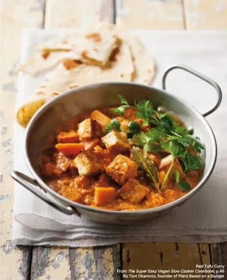 A metal bowl of red tofu curry with flatbread on a rustic table.