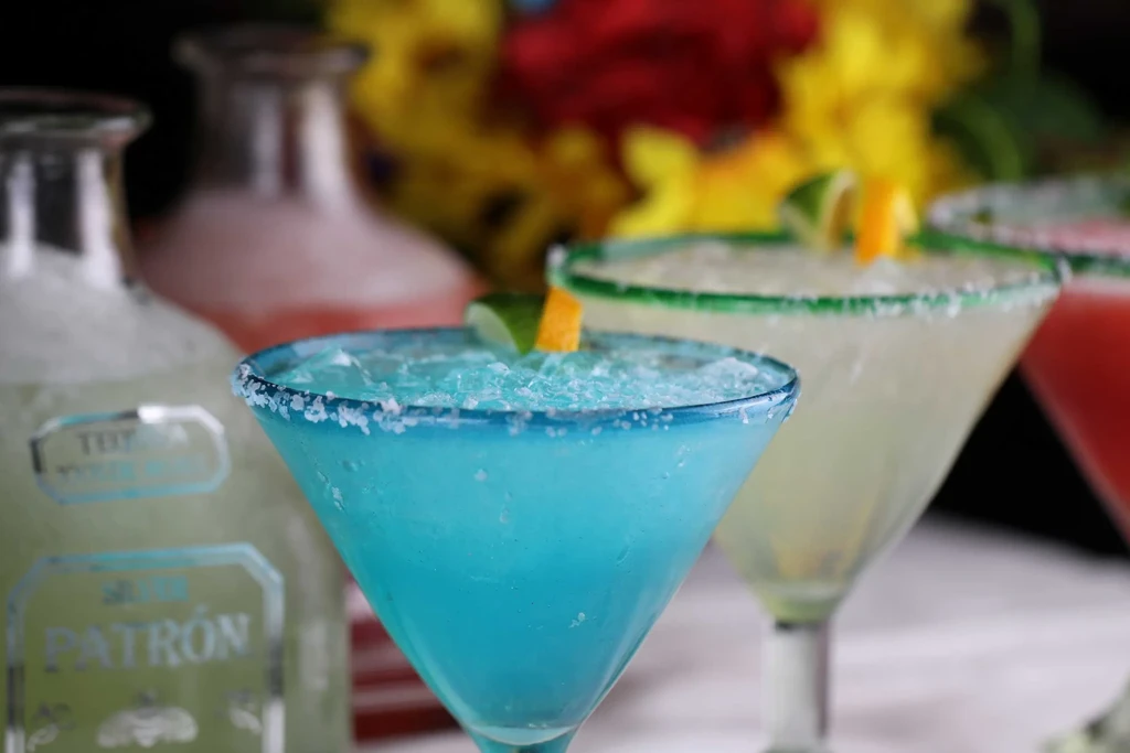 Three frozen margaritas, with tequila bottles and flowers in the background.