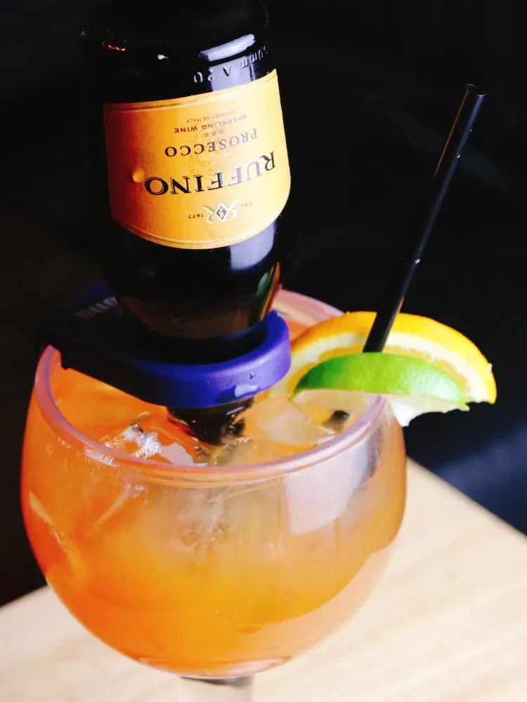 Miniature prosecco bottle upside down in an orange cocktail with citrus garnish.