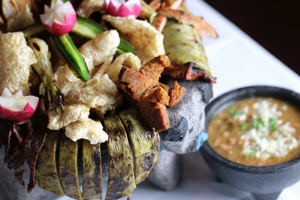 Molcajete bowl filled with meat, pork rinds, grilled vegetables, and radishes.