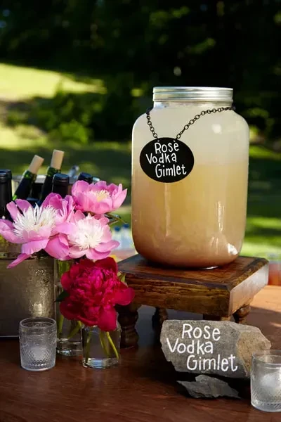 A large jar of Rose Vodka Gimlet on a wooden stand outdoors.