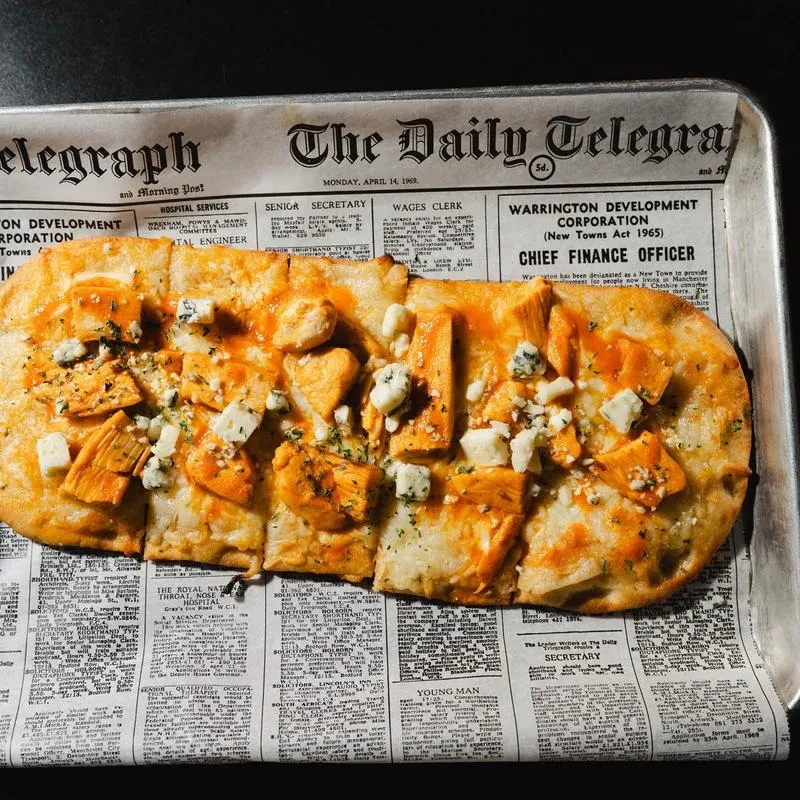 Buffalo chicken flatbread with blue cheese, served on newspaper-lined metal tray.