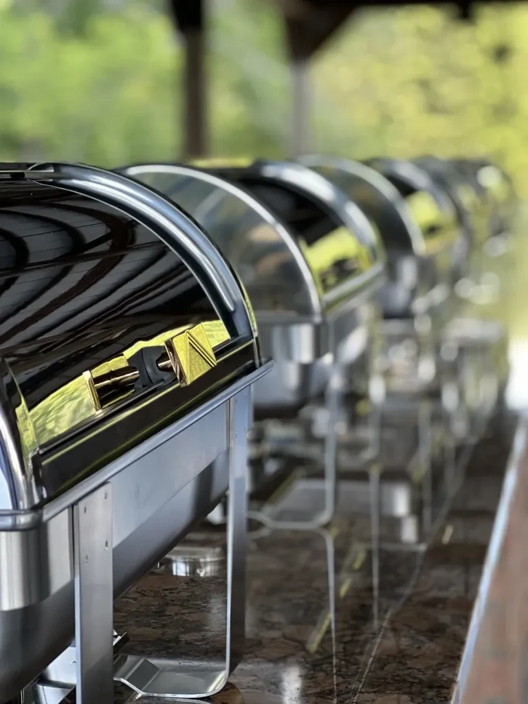 A row of stainless steel chafing dishes on a reflective counter with blurred foliage.