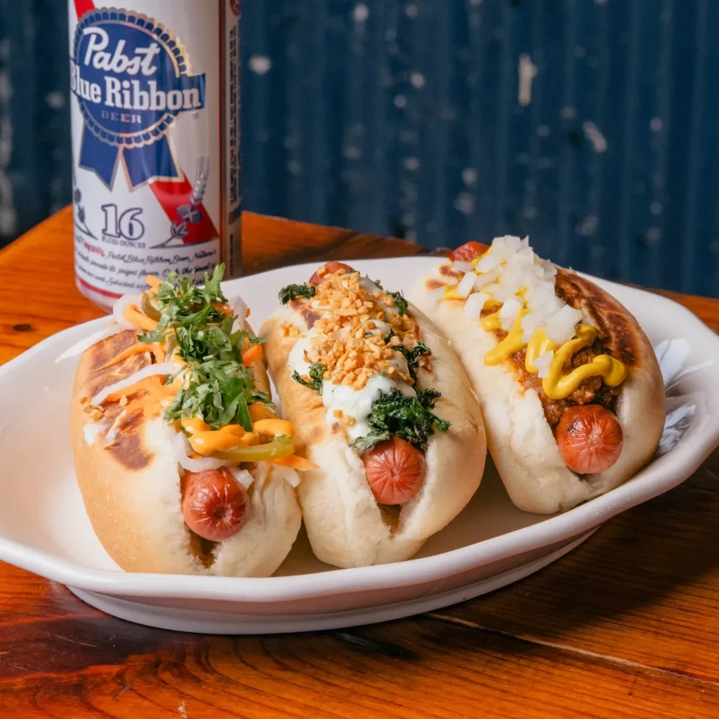 Three gourmet hot dogs with distinct toppings, plate, and PBR can.