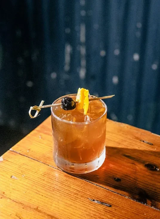 Amber cocktail with orange peel and cherry on a rustic table.