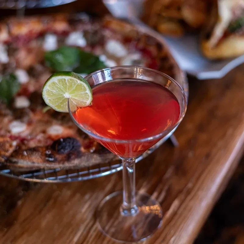 Red cocktail with lime garnish; blurred pizza in restaurant background.
