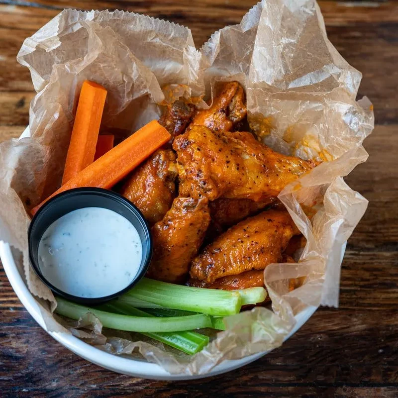 White bowl of buffalo wings, carrots, celery, and dip on wood.