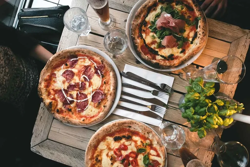 Overhead of three pizzas, drinks, and cutlery on a rustic table.