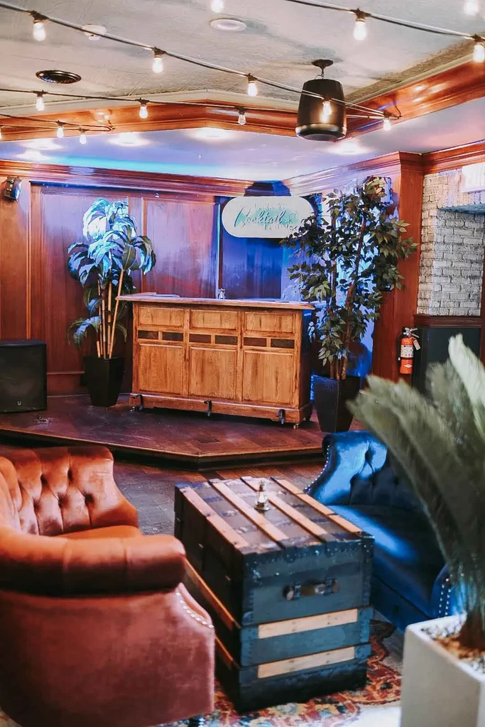 Bar lounge with armchairs, stacked trunks, and an illuminated 'Cocktails' sign.