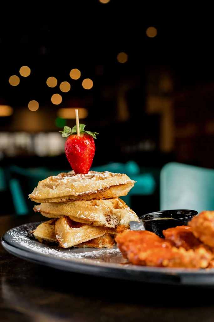 Stacked golden waffles with strawberry, sauced chicken, on a dark plate.