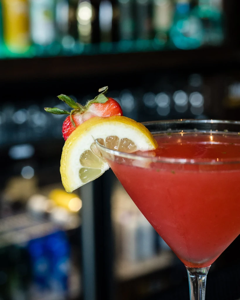 Red cocktail in martini glass, garnished with strawberry and lemon.