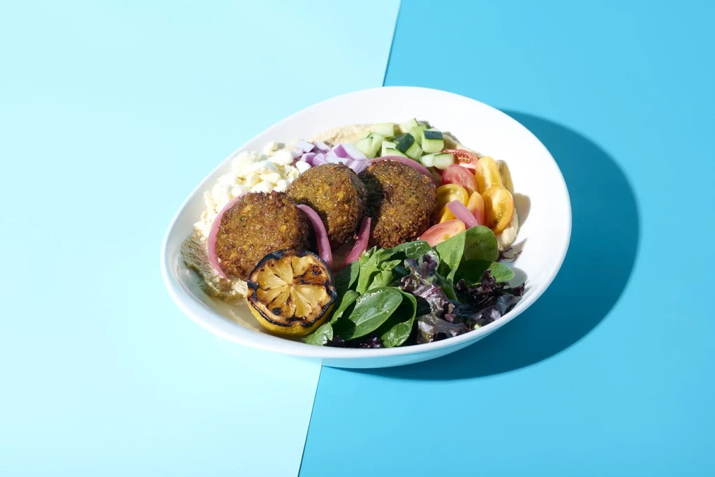 Oval bowl with falafel, feta, vegetables, and grilled lemon on blue.