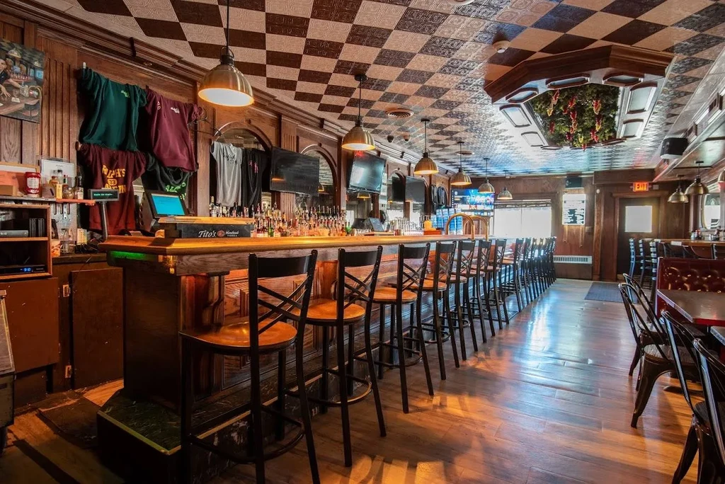Rustic bar interior with a long wooden counter, stools, and liquor.