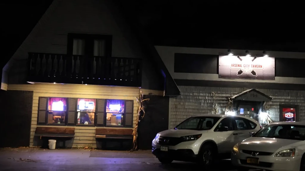 Arsenal City Tavern at night; illuminated sign, balcony, parked cars.