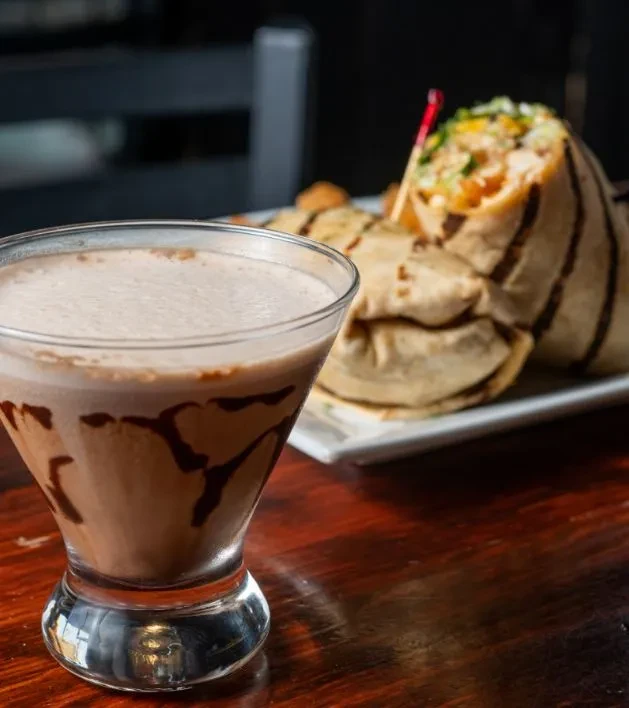 Frothy chocolate drink in martini glass and a burrito on wood.