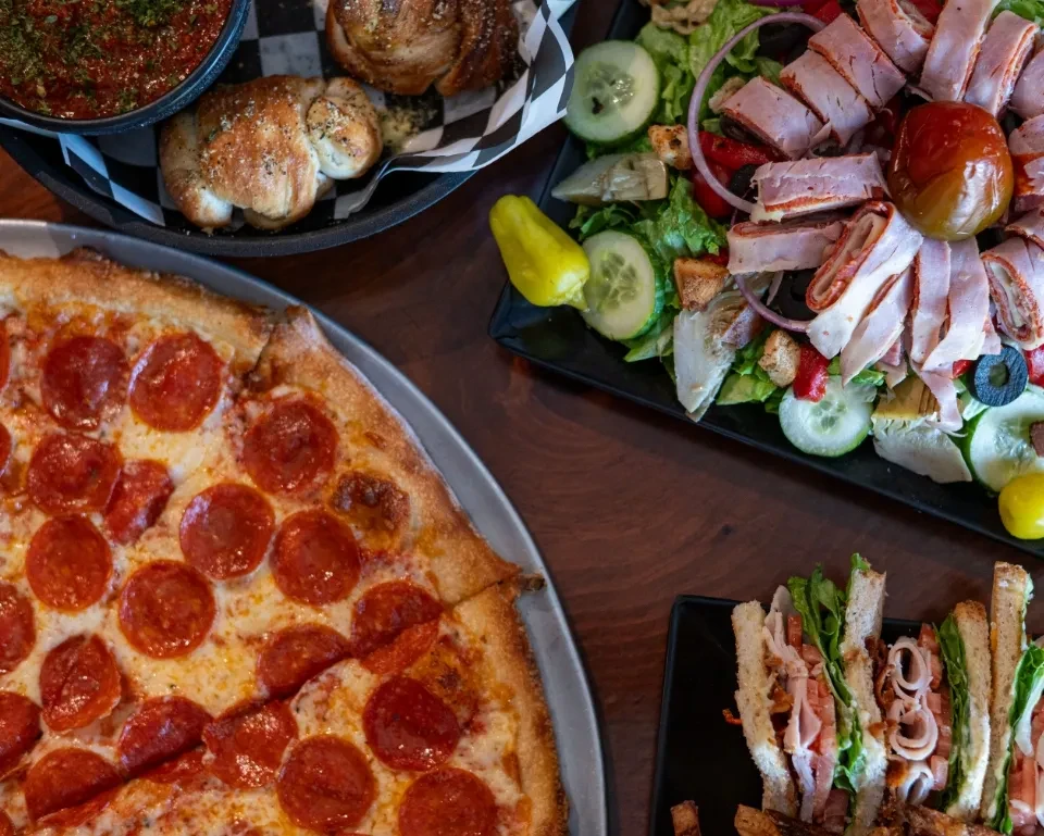 Overhead view of pepperoni pizza, garlic knots, antipasto salad, and club sandwiches.