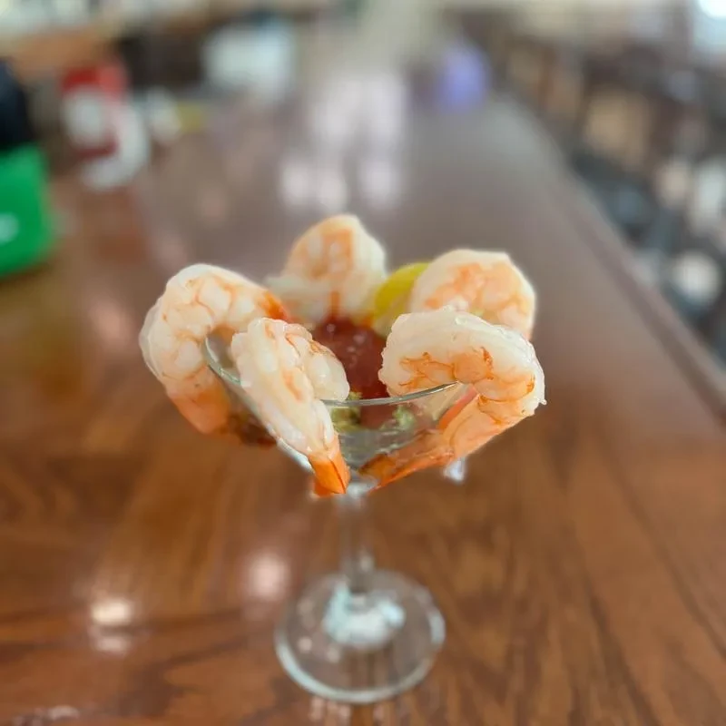 Cooked shrimp cocktail with sauce and lemon in a stemmed glass on wood.