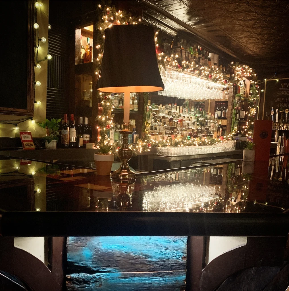 Warmly lit bar interior with reflective counter, stocked bottles, and fairy lights.