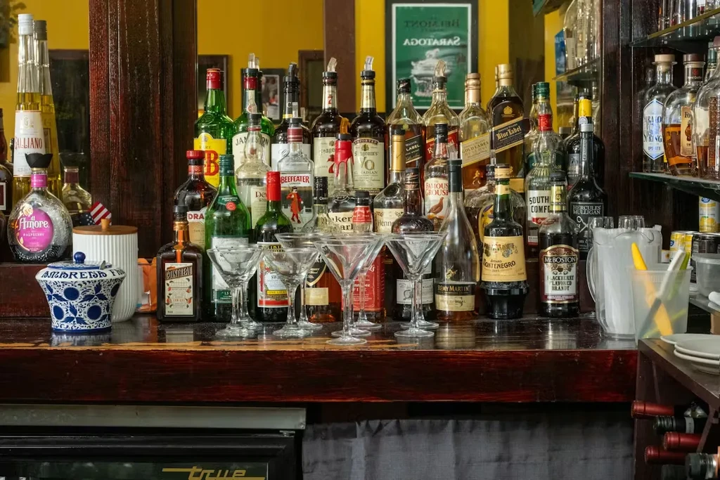 Well-stocked bar counter with diverse liquor bottles, empty martini glasses, and tools.