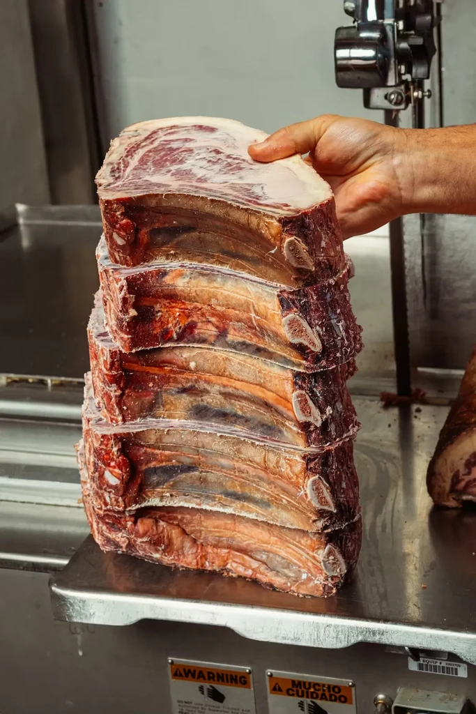 A hand holds five stacked raw, marbled bone-in ribeye cuts.