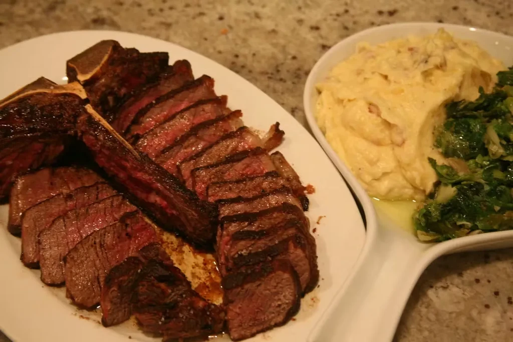 Sliced bone-in steak, mashed potatoes, and leafy green vegetables.