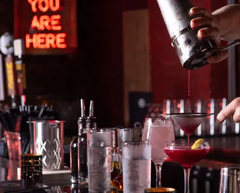 Bartender's hands strain a crimson cocktail into a martini glass.