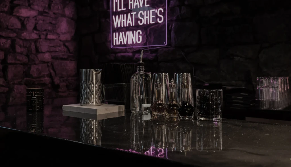 Dimly lit bar counter with barware and purple neon sign.