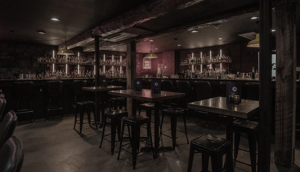Dimly lit bar with backlit bottles and a "YOU ARE HERE" neon sign.