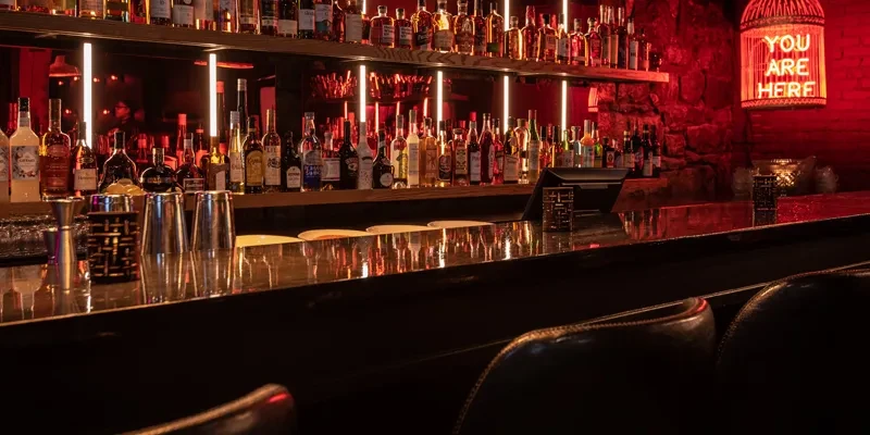 Red-lit bar, backlit bottles, bar tools, 'YOU ARE HERE' neon sign.