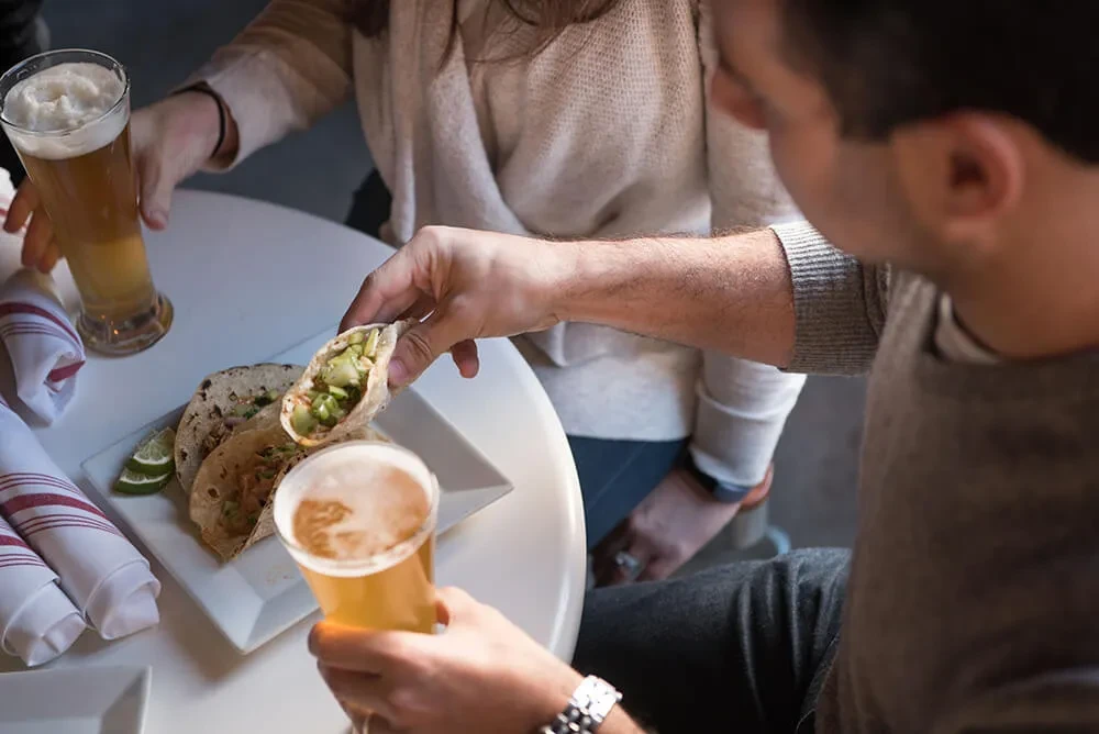 Overhead view of hands holding tacos and beer glasses at a table.