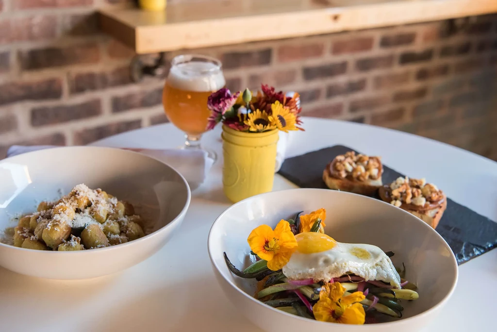 Table with gnocchi, green beans, pastries, beer, and colorful flowers.
