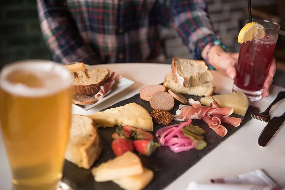 Overhead charcuterie board with cheeses, meats, bread, strawberries, beer, and cocktail.