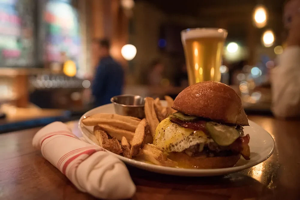 Burger with egg, fries, and beer on a wooden restaurant table.