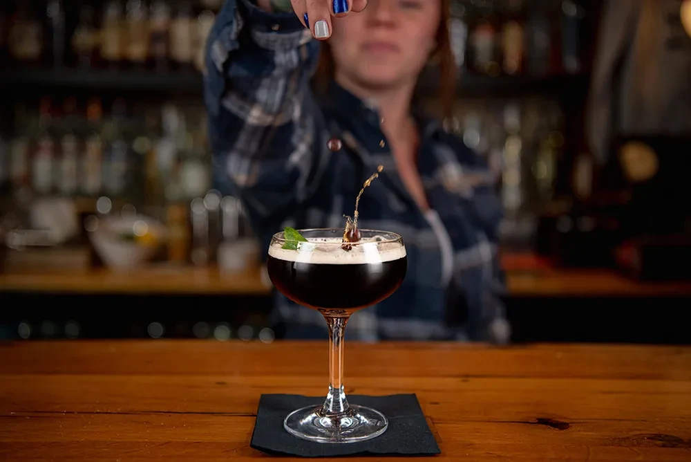 Dark cocktail with foam and mint splashes as bartender garnishes it.